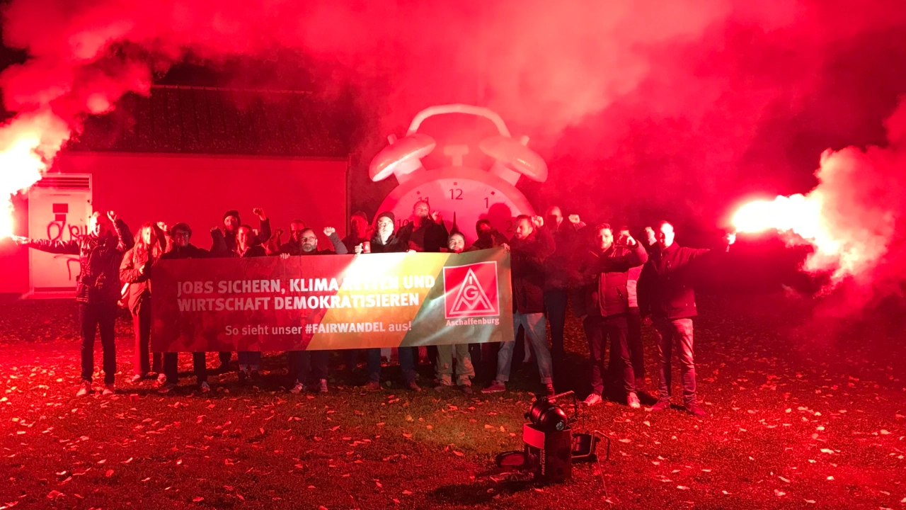 Das Bild zeigt Aktive der IG Metall Aschaffenburg mit einem Banner mit der Aufschrift: Jobs sicher, Klima retten, Wirtschaft Demokratisieren - So sieht unser #Fairwandel aus! Umrahmt wird die Szene von einem großen Wecker und roten Bengalo-Fackeln