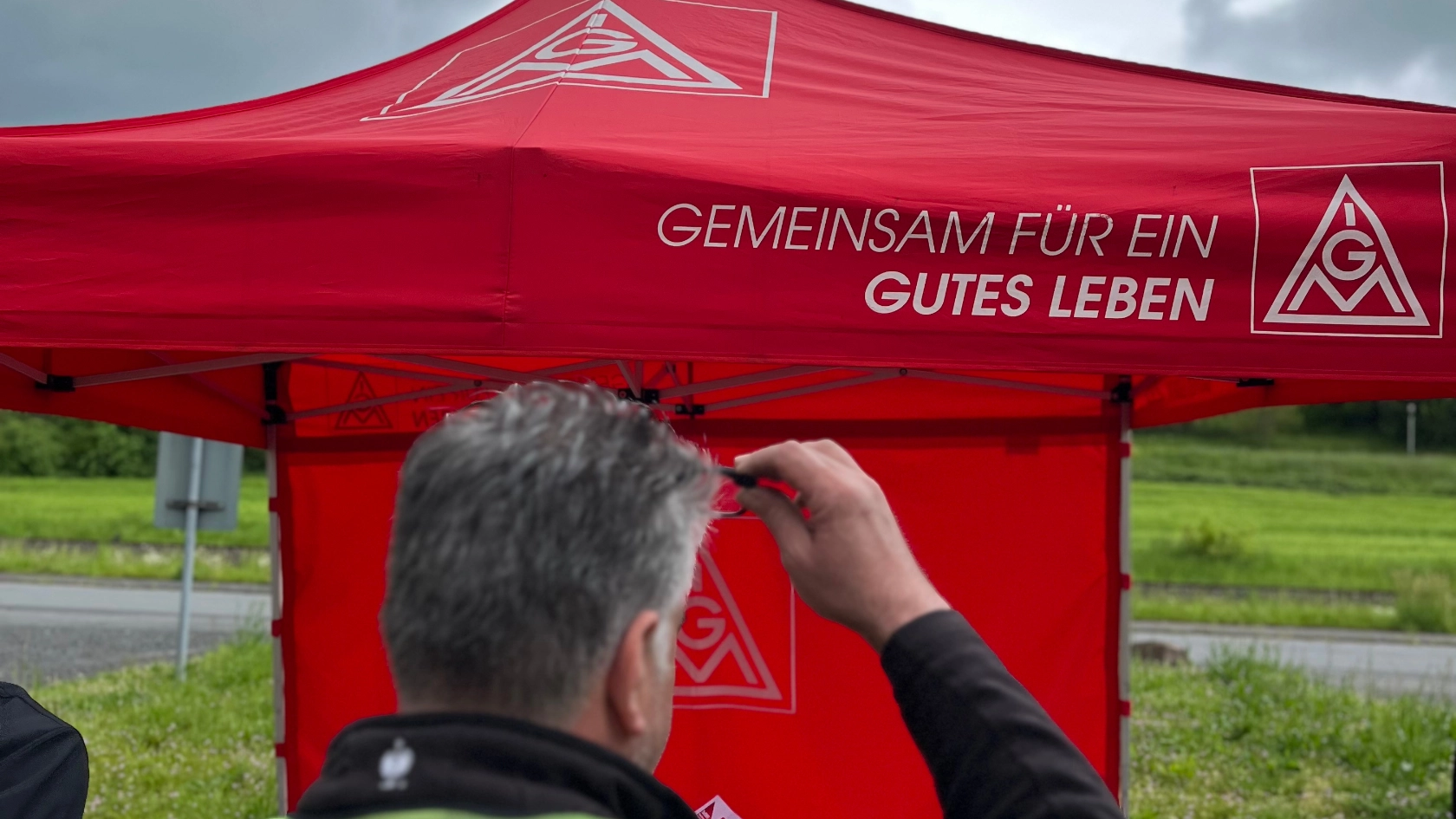 Ein Mitarbeiter mit MAGNA Weste steht vor den IG Metall Pavillon mit der Aufschrift: Gemeinsam für ein gutes Leben