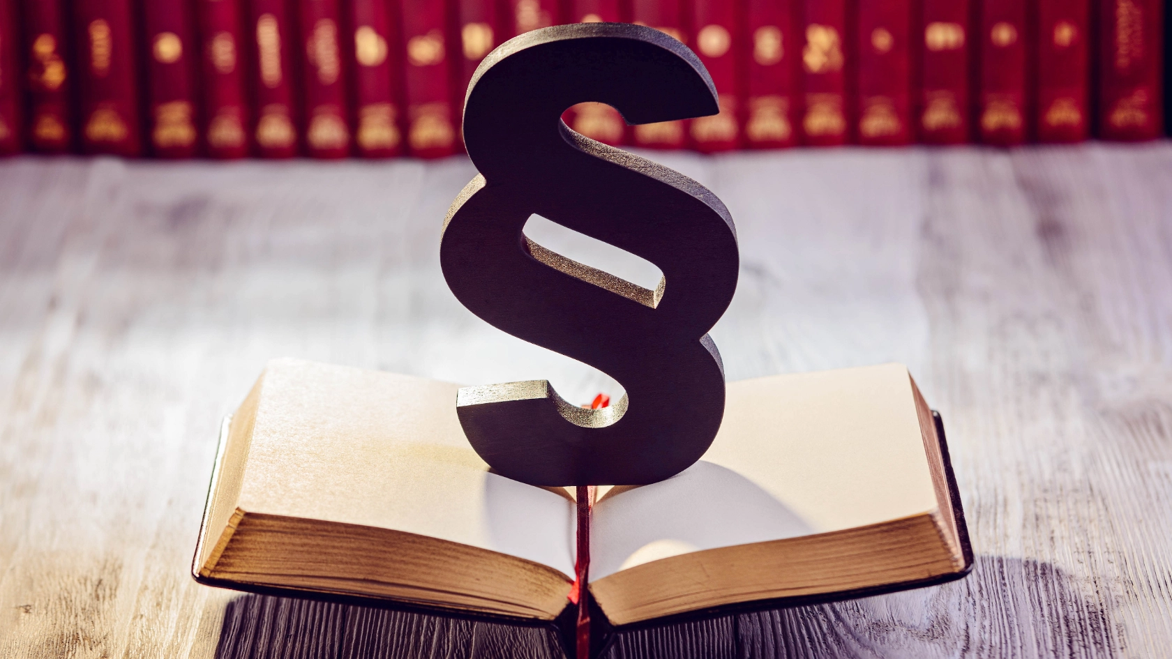 Ein schwarzer Paragraph steht auf einem geöffneten Gesetzbuch, im Hintergrund sind unscharf weitere Bücher zu sehen