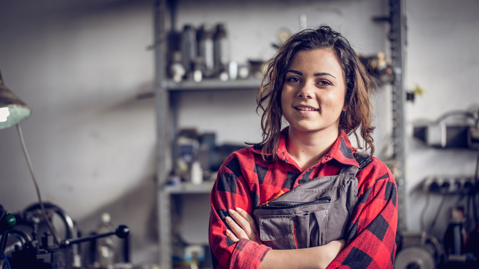 Junge Mechanikerin mit rotkariertem Hemd und Latzhose schaut mit verschränkten Armen selbstbewusst in die Kamera.