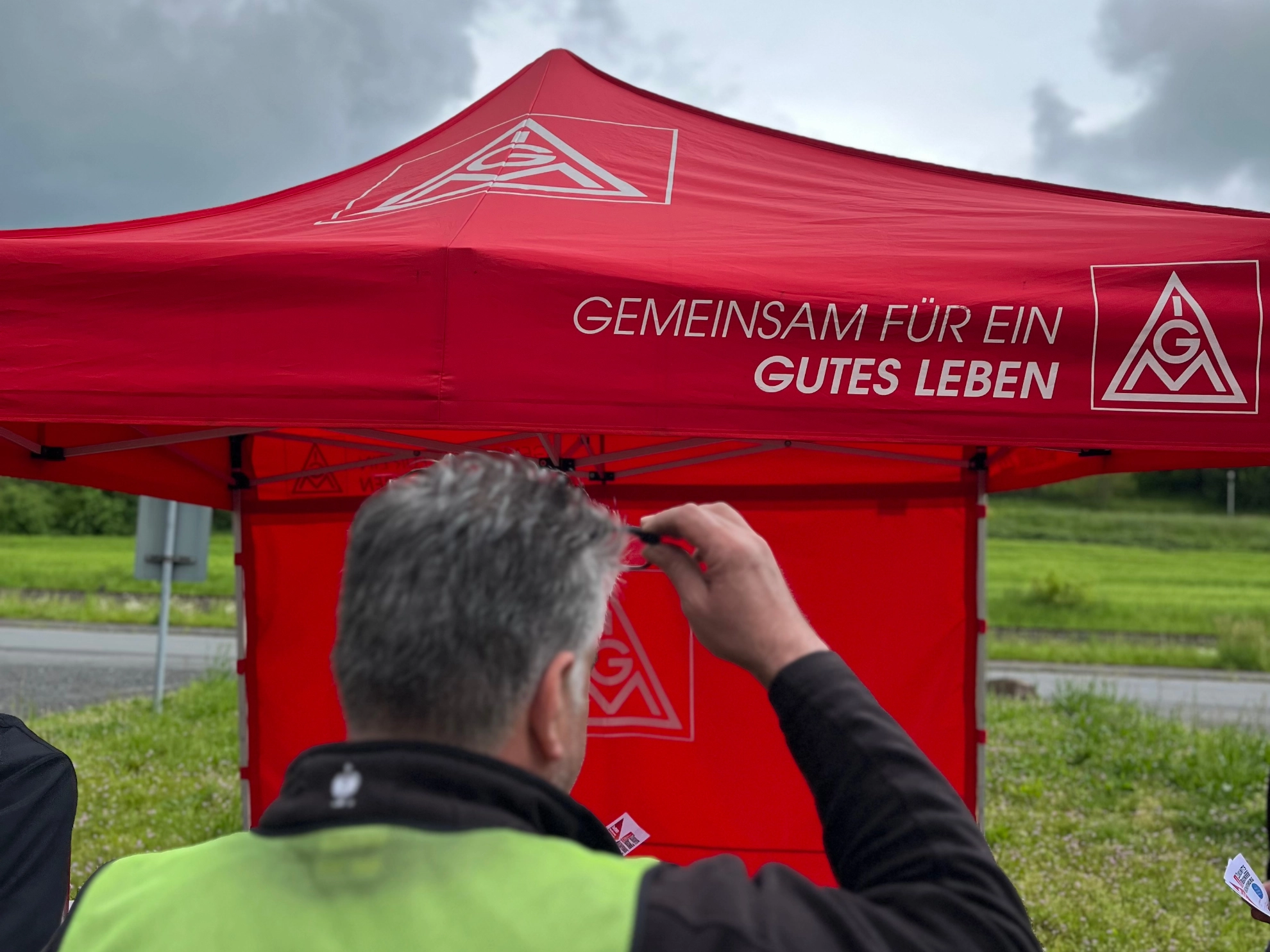Ein Mitarbeiter mit MAGNA Weste steht vor den IG Metall Pavillon mit der Aufschrift: Gemeinsam für ein gutes Leben