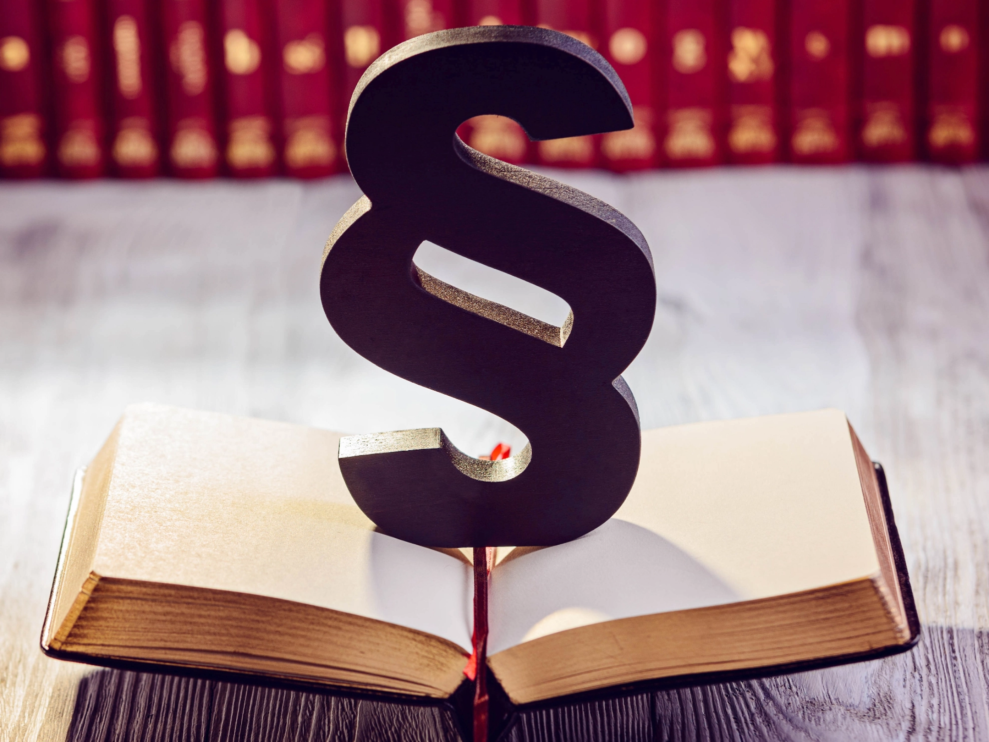 Ein schwarzer Paragraph steht auf einem geöffneten Gesetzbuch, im Hintergrund sind unscharf weitere Bücher zu sehen
