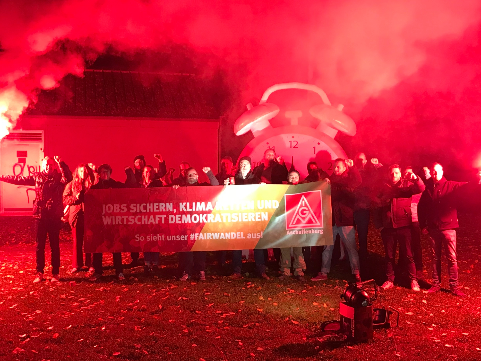 Das Bild zeigt Aktive der IG Metall Aschaffenburg mit einem Banner mit der Aufschrift: Jobs sicher, Klima retten, Wirtschaft Demokratisieren - So sieht unser #Fairwandel aus! Umrahmt wird die Szene von einem großen Wecker und roten Bengalo-Fackeln