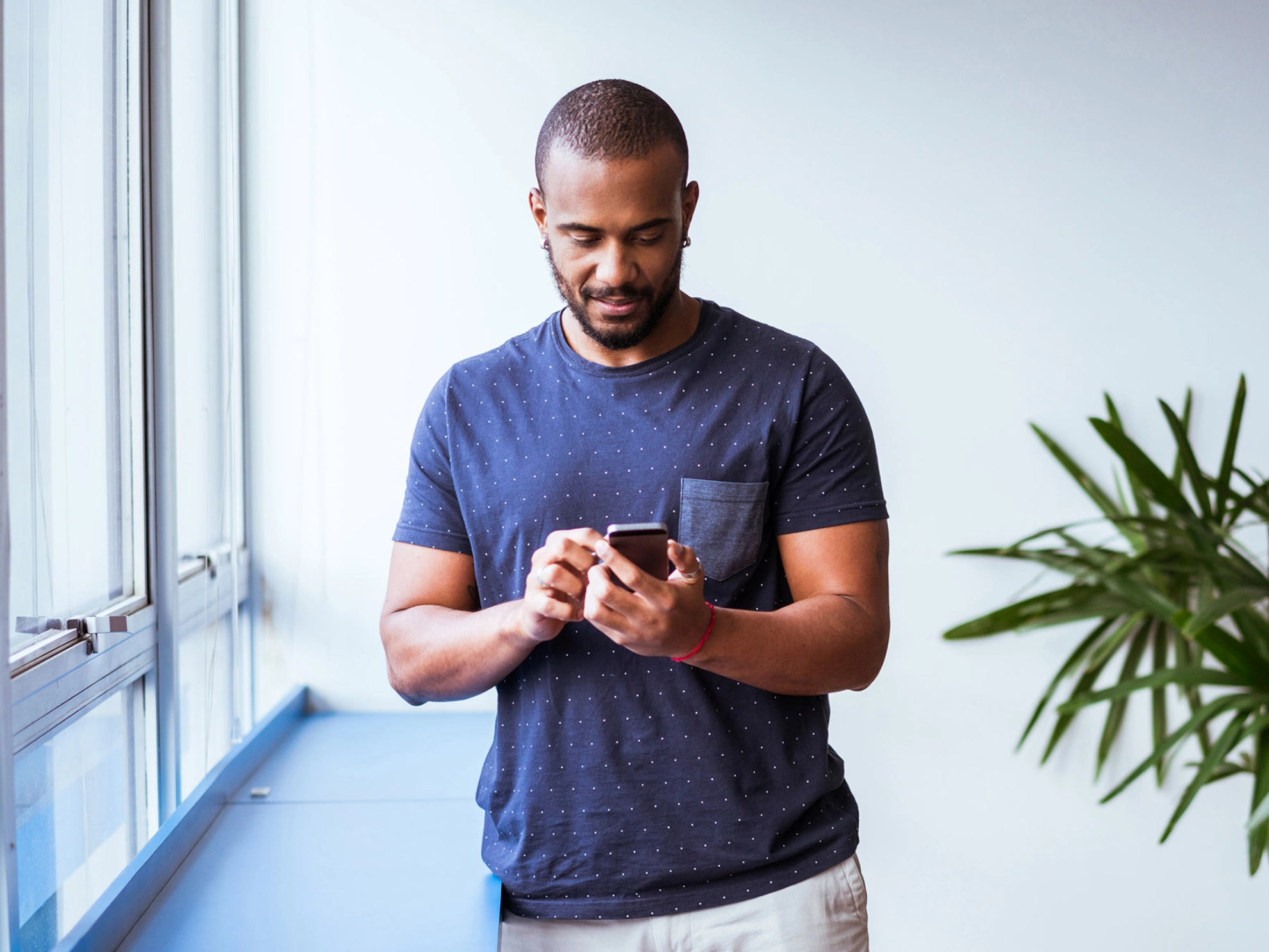 Ein dunkelhaariger man steht vor einem Fesnster und tippt auf seinem Smartphone.
