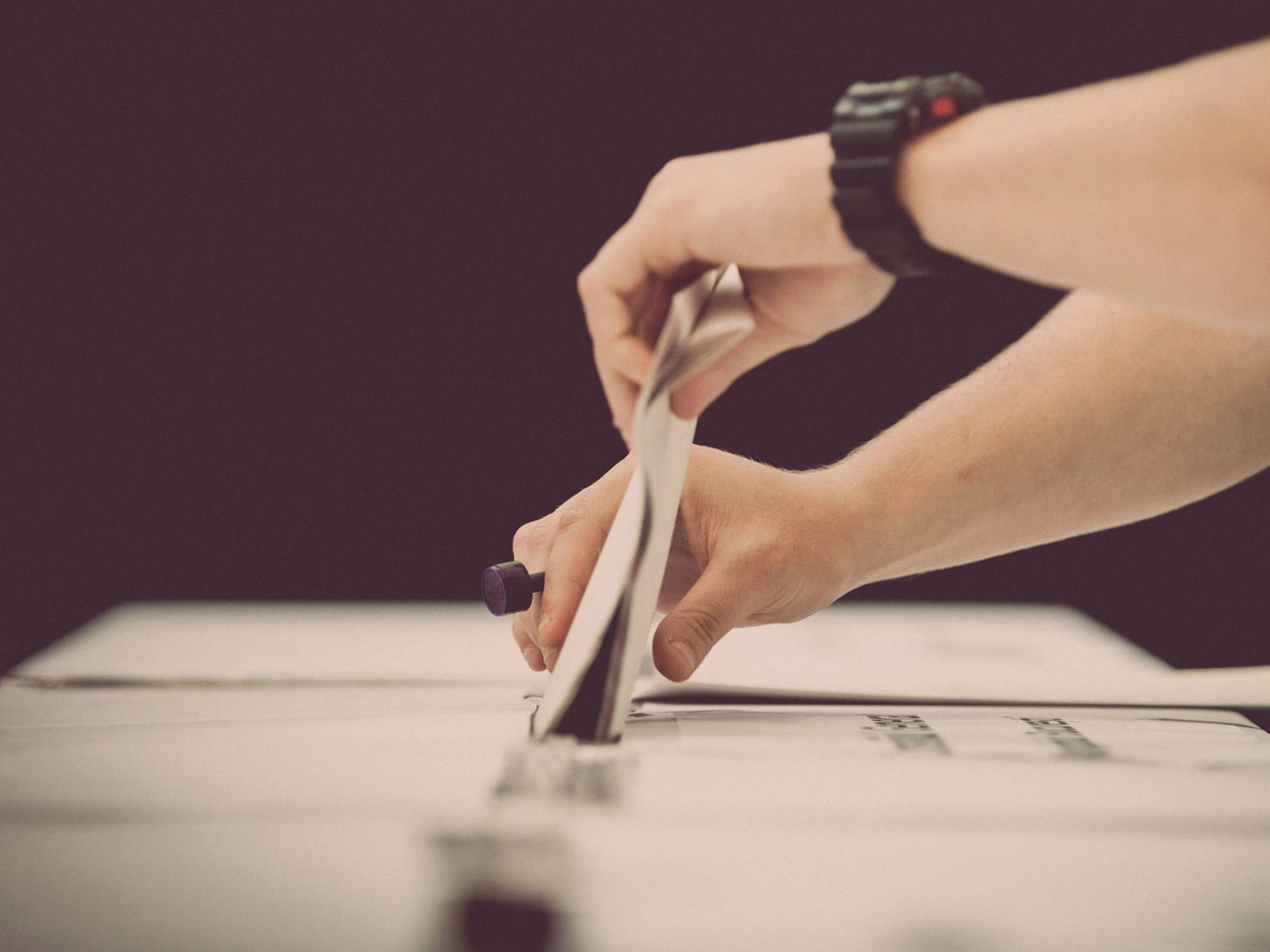 Jemand mit einer schwarzen Sportuhr am linken Handgelenkt steckt mit beiden Händen einen Stimmzettel in den Schlitz einer Wahlurne.