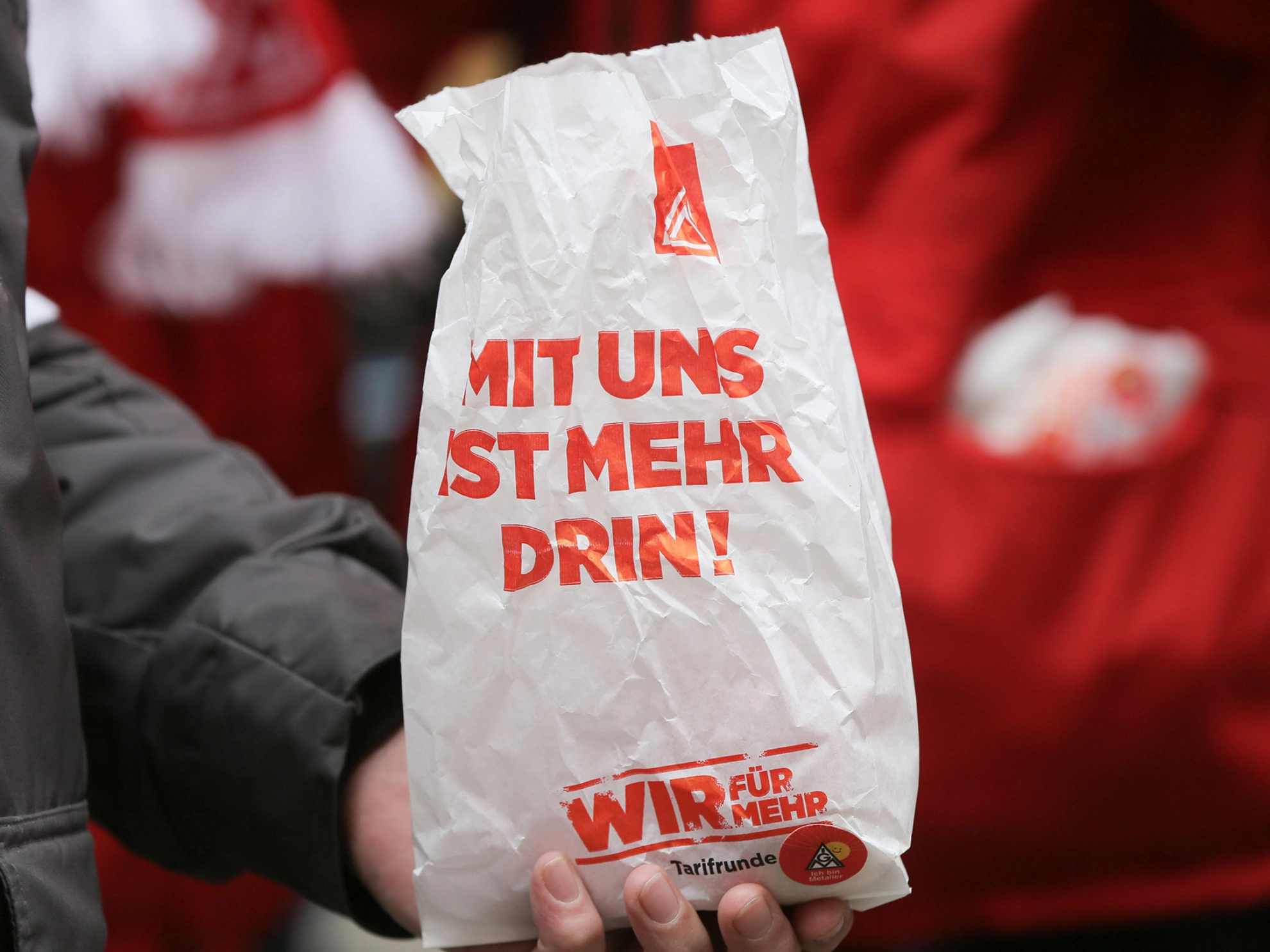 Hand hält Papiertüte mit "Mit uns ist mehr drin!"-Slogan.